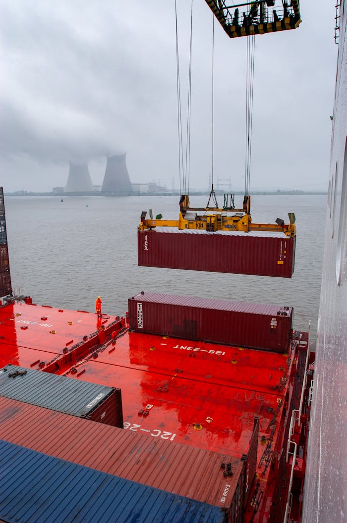 A crane lifts a shipping container on a rainy day at an industrial port with cooling towers in the background.